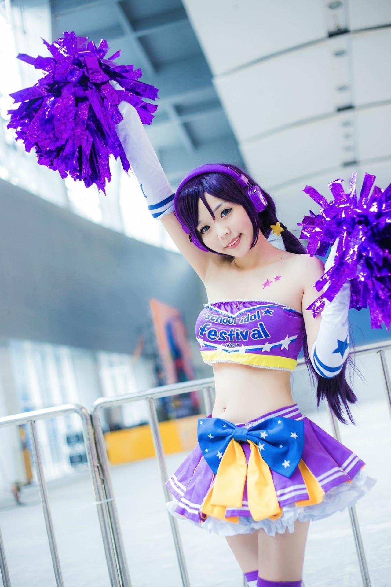 蓝天盈盈子扮演东条希右手举起花球，左手将花球举到肩部，背景是栏杆