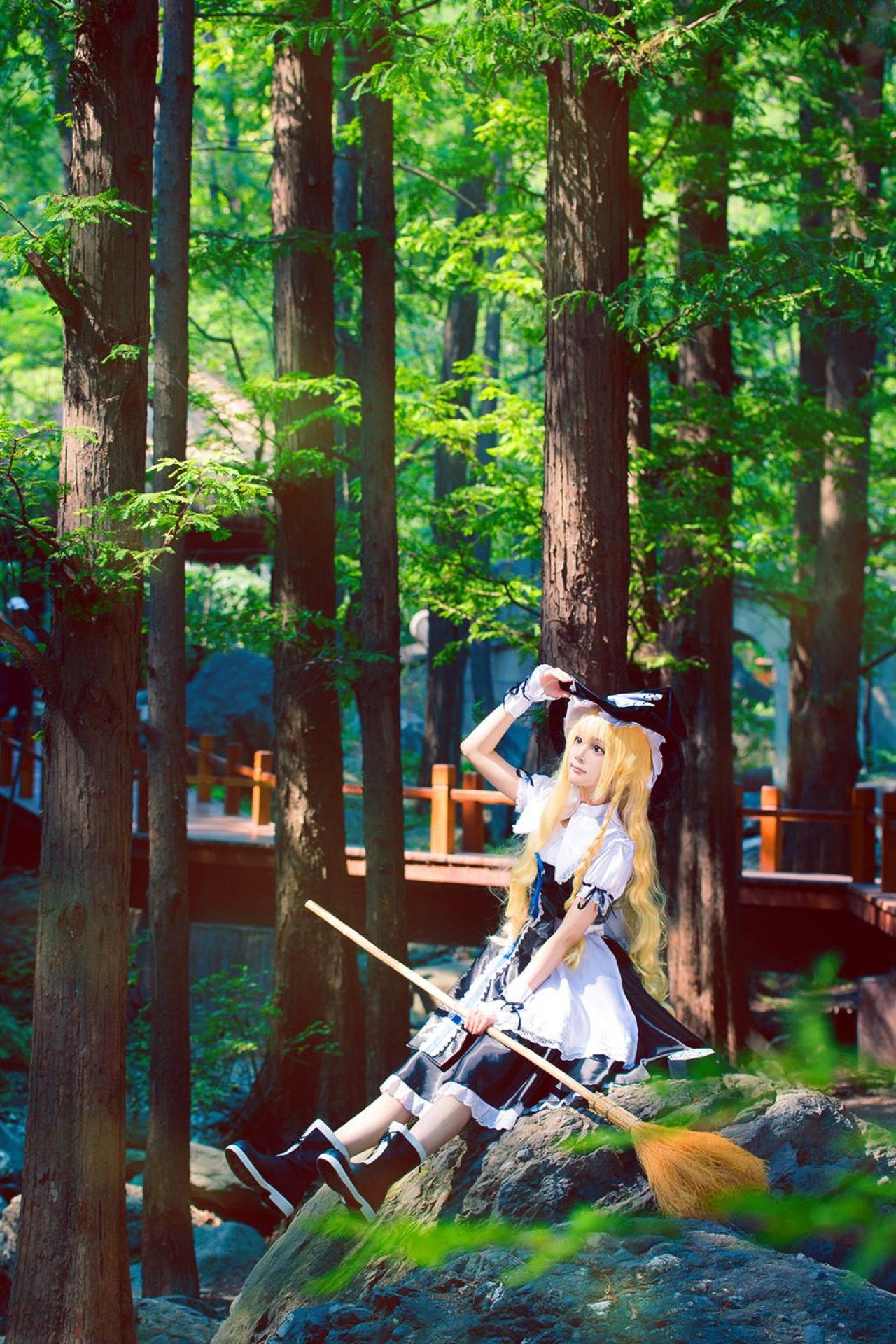 雨扮演雾雨魔理沙，右手扶着帽檐，左手拿着扫把坐在水边