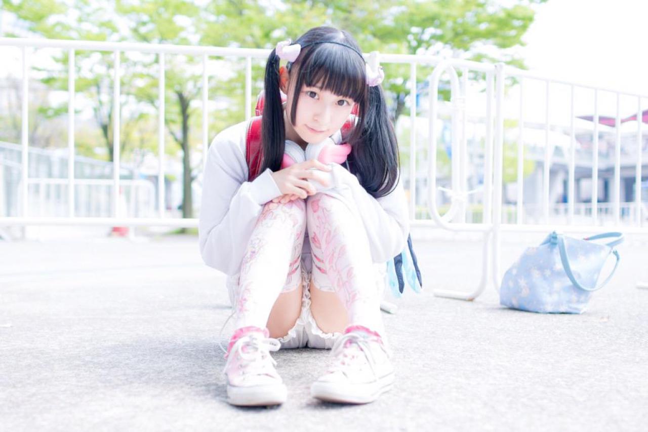 猫守ざーにゃ怀抱双腿，双手五指相抵搭在膝盖上，坐在栏杆前，好奇地看着镜头