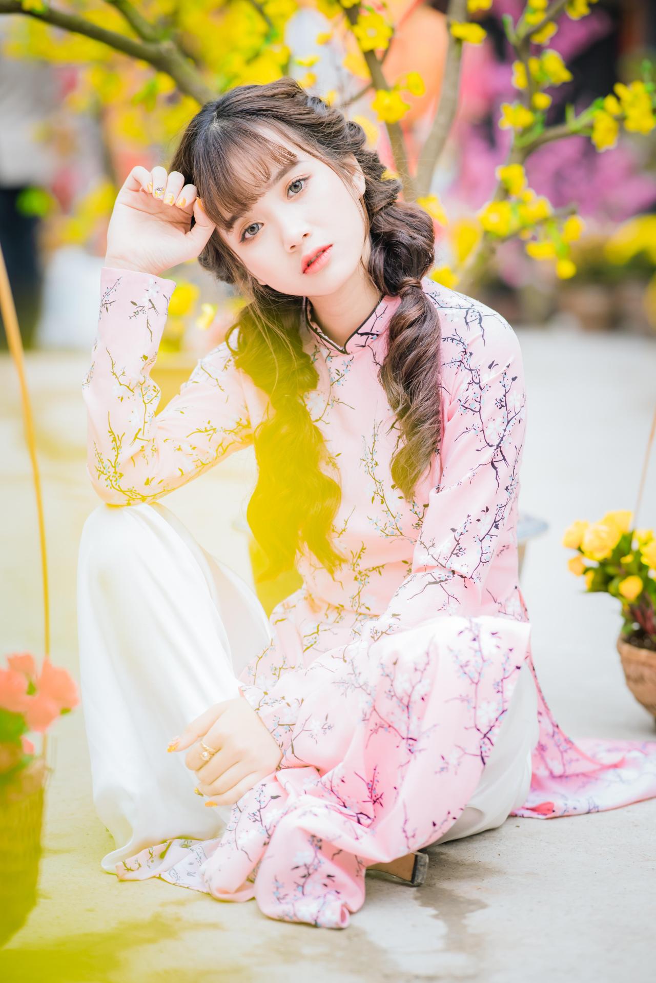 棕色双马尾卷发，齐刘海儿，穿着粉色旗袍白色长裤的少女 坐姿 全身照 4/3侧面照 竖幅 咧嘴露齿微笑 右手轻抚额头 腊梅 黄梅 花篮