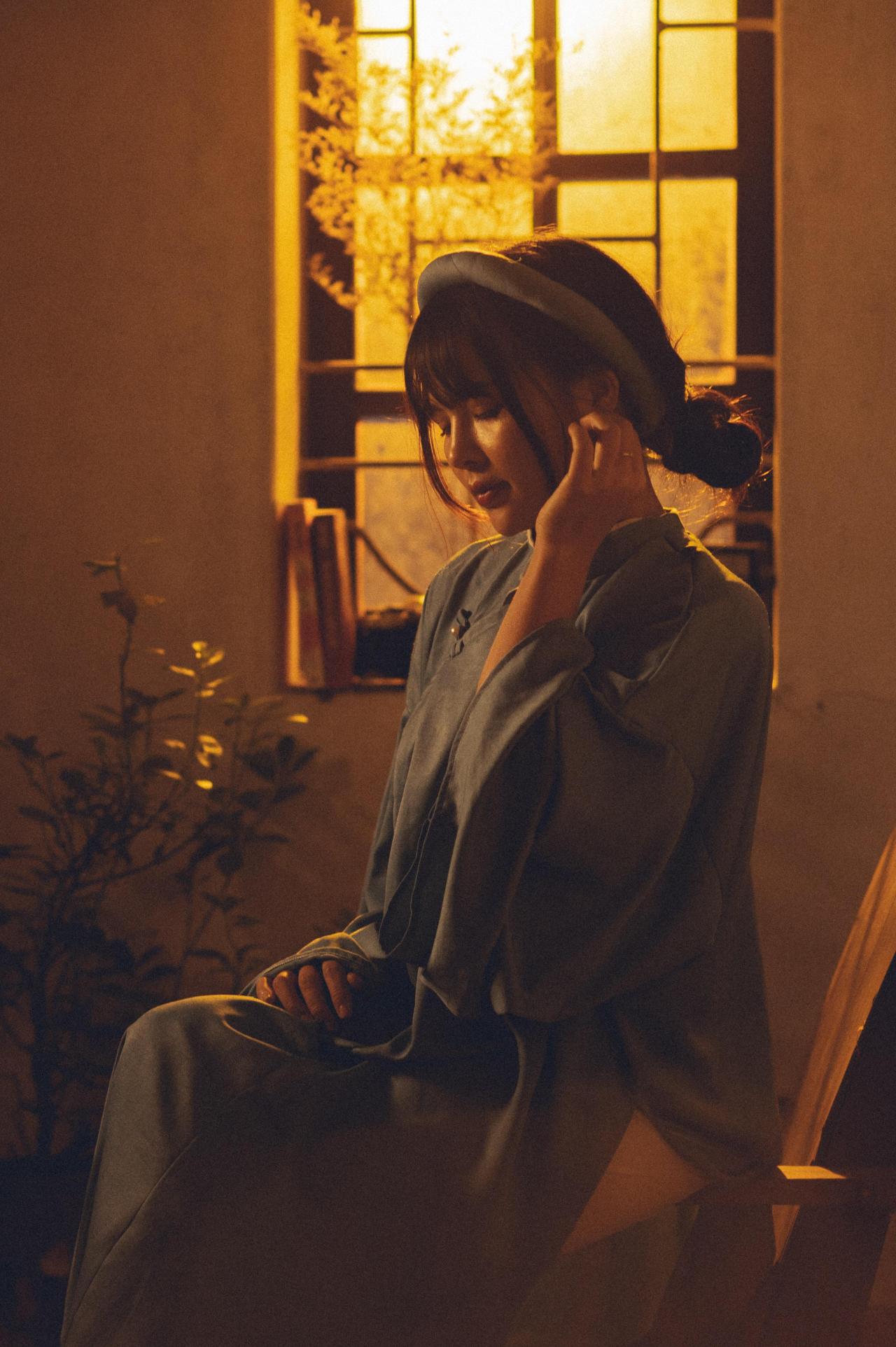 黄昏老屋窗边穿着蓝色越南长袄奥黛少女靠坐在椅子的扶手上，戴着蓝色发箍，轻抚耳垂，4/3 斜侧面照 竖幅 全身照